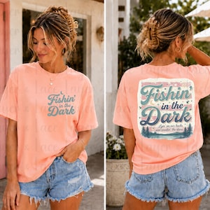 May include: A peach-colored short-sleeve t-shirt featuring the text Fishin' in the Dark on the front left chest. The back displays a larger graphic with the same text, a moon, stars, and a forest landscape with the phrase Lyin' on our backs and countin' the stars.