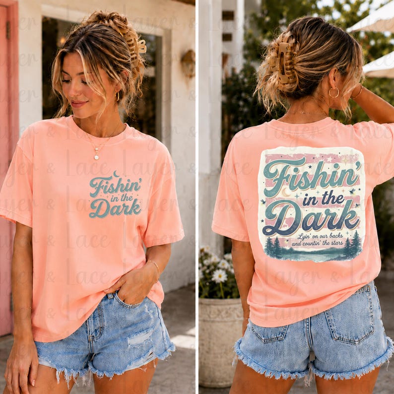 May include: A peach-colored short-sleeve t-shirt featuring the text Fishin' in the Dark on the front left chest. The back displays a larger graphic with the same text, a moon, stars, and a forest landscape with the phrase Lyin' on our backs and countin' the stars.