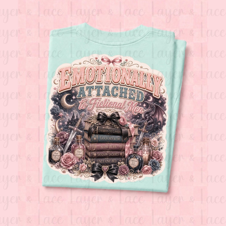May include: A light blue t-shirt featuring the text "Emotionally Attached to Fictional Men" in a decorative font. The design includes a stack of books, roses, swords, and a dragon, creating a fantasy theme.