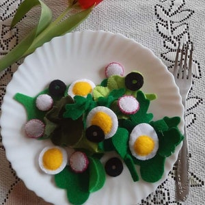 Op de afbeelding: Een set met nepvoedsel in de vorm van een salade op een wit bord met een vork. De salade bevat groene sla, witte en rode radijsjes, zwarte olijven en gele eieren.