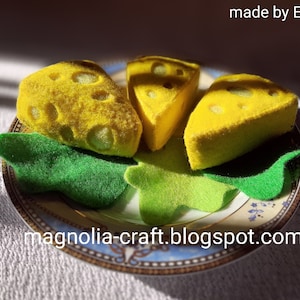 Pode incluir: Três fatias de queijo de feltro em um prato decorativo com folhas de feltro verde. O queijo é amarelo com furos e as folhas são verdes. O prato tem um design azul e dourado. O texto "made by EDITH" e "magnolia-craft.blogspot.com" são visíveis na imagem.