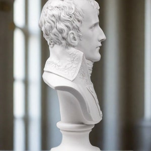 May include: White plaster bust sculpture of a man with short hair, wearing a collared shirt. The bust is on a white pedestal.