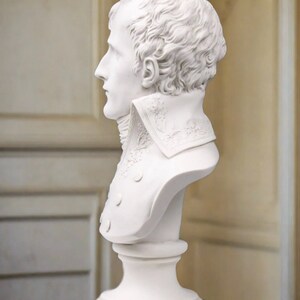 May include: White plaster bust sculpture of a man with short, wavy hair. The bust is on a white pedestal.