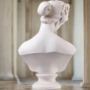 May include: White marble bust sculpture of a woman with her hair pulled back in a bun. The bust is on a pedestal.
