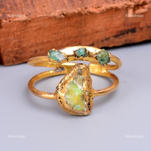 Op de afbeelding: Een gouden ring met een grote, onregelmatig gevormde groene edelsteen en drie kleinere groene edelstenen. De ring is gemaakt van twee banden, waarvan er één gebogen is.