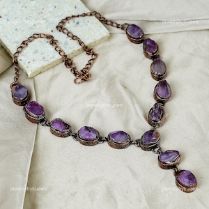 Pode incluir: Um colar de corrente de cobre com um pingente feito de várias pedras de ametista. As pedras são de cor roxo escuro e têm uma textura áspera e natural.