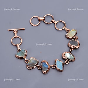 Puede incluir: Una pulsera de oro rosa con piedras de labradorita de forma irregular. Las piedras muestran tonos iridiscentes azules, verdes y grises. La pulsera tiene un cierre de palanca y está etiquetada como "jewelrybykuen".