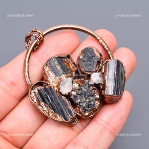 May include: A copper wire wrapped pendant featuring a cluster of black tourmaline and clear quartz crystals. The crystals are set in a copper frame and are surrounded by a copper wire loop.