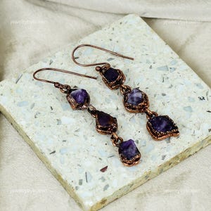 Może przedstawiać: Para kolczyków z miedzi z kamieniami ametystowymi. Każdy kolczyk ma cztery ametysty o różnych rozmiarach i kształtach, połączone miedzianym drutem. Kolczyki mają prosty, elegancki design.