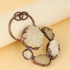 May include: A copper-coloured pendant with a circular design. It features sections of light-coloured, crystalline stones set in a copper-coloured metal frame. A twisted loop at the top allows for hanging.
