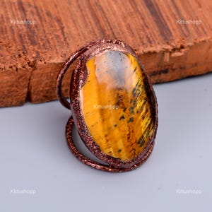 May include: A copper wire wrapped ring with a large oval shaped tiger's eye gemstone. The gemstone is a golden yellow color with brown and black stripes.
