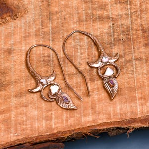 May include: A pair of copper-colored earrings with a floral design. Each earring features a curved hook, a flower-like top, a white stone, and a leaf-shaped charm with a purple stone. The earrings are displayed on a wooden surface.