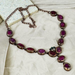 Könnte beinhalten: Eine Kupferkette mit einem Anhänger, der aus einer Gruppe von dunkelvioletten Edelsteinen besteht. Die Kette hat eine Kette mit einer Reihe von ovalen dunkelvioletten Edelsteinen.