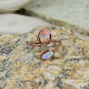 May include: A rose gold wire wrapped ring with three opal stones. The ring is designed with a spiral wire and features a larger round opal stone and two smaller opal stones.