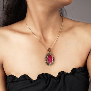 May include: A gold-toned necklace featuring an oval pendant with a large, vibrant red gemstone at its center. The pendant is framed by smaller green and clear stones, and the necklace has a delicate chain. The overall design is elegant and eye-catching.