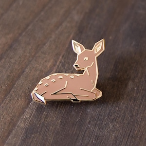 May include: A brown enamel pin depicting a stylized deer in a resting pose. The pin features a gold outline and small white spots. It is set against a dark wood grain background.