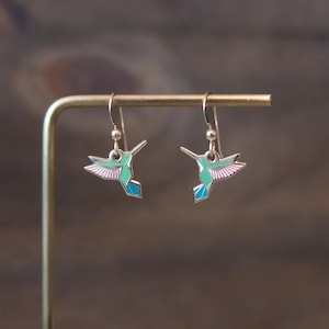 May include: A pair of hummingbird earrings. Each earring features a silver-colored hook and a hummingbird charm. The hummingbird charms are teal, pink, and blue, with outstretched wings. The earrings are displayed on a gold-colored stand.