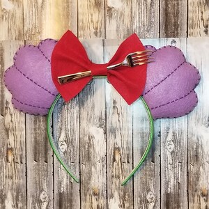 May include: A headband featuring purple felt seashell shapes with stitched details, a red bow, and a silver fork accent. The headband has a green band. The design is inspired by a popular animated film.