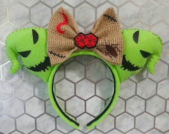 May include: A lime green headband with two plush monster ears. A burlap bow features a red dice design, a red flame, and a brown insect. The headband is black and green, ideal for Halloween or themed events.