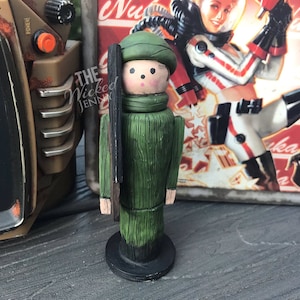 May include: A wooden figurine of a soldier in a green uniform, hat, and scarf, holding a rifle. The figurine has a painted face and is standing on a black base. The Wicked Jenny is written on the side.