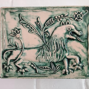 May include: A rectangular ceramic tile with a relief design of a mythical creature with wings, a lion's body, and a dog-like head. The tile is primarily light pink with green accents and a green border. Floral elements are also present.