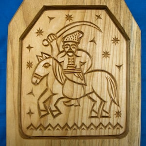 May include: Wooden plaque featuring a carved relief of a figure on horseback wielding a sword. The figure wears a hat and mustache, surrounded by stars. The plaque has a decorative border and is made of light-colored wood.