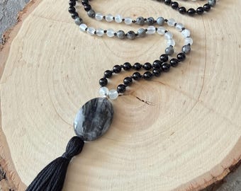 Può includere: Una collana di perline nere con un grande pendente ovale e un nappa nera. La collana presenta perline nere, grigie e bianche alternate. Il pendente è una pietra grigia con venature nere.