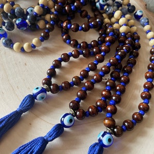May include: Multiple strands of wooden bead necklaces. The beads are brown, cream, and blue. Each necklace features a blue tassel and an evil eye bead. The necklaces are displayed on a light wood surface.