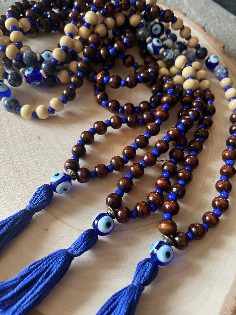 May include: Multiple strands of wooden bead necklaces. The beads are brown, cream, and blue. Each necklace features a blue tassel and an evil eye bead. The necklaces are displayed on a light wood surface.