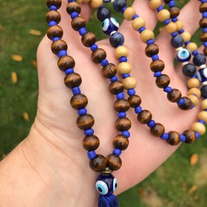 May include: A beaded necklace with alternating dark brown and light wood beads, linked by blue beads. The necklace features blue evil eye charms and a blue tassel. The beads are strung on a blue cord.