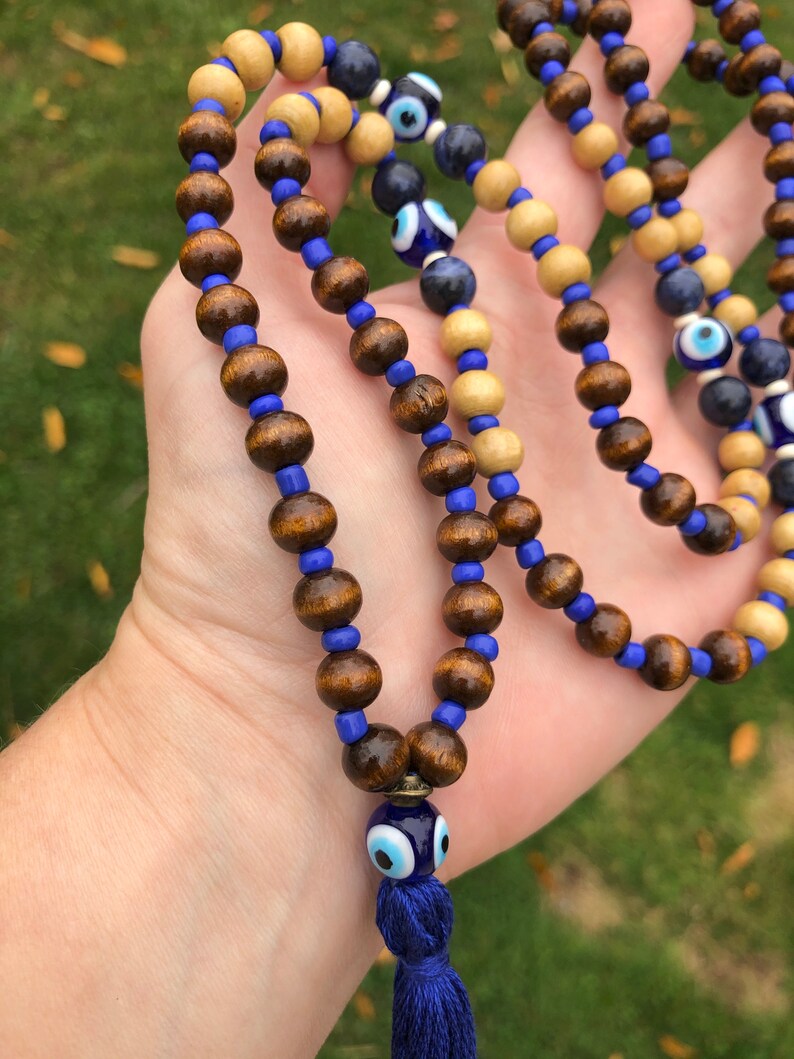 May include: A beaded necklace with alternating dark brown and light wood beads, linked by blue beads. The necklace features blue evil eye charms and a blue tassel. The beads are strung on a blue cord.