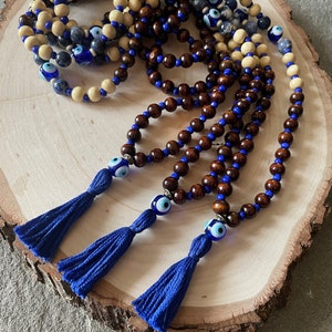 May include: A collection of beaded necklaces featuring wooden beads in brown and cream, with blue accents and tassels. Each necklace has an evil eye charm. The necklaces are presented on a wooden surface.