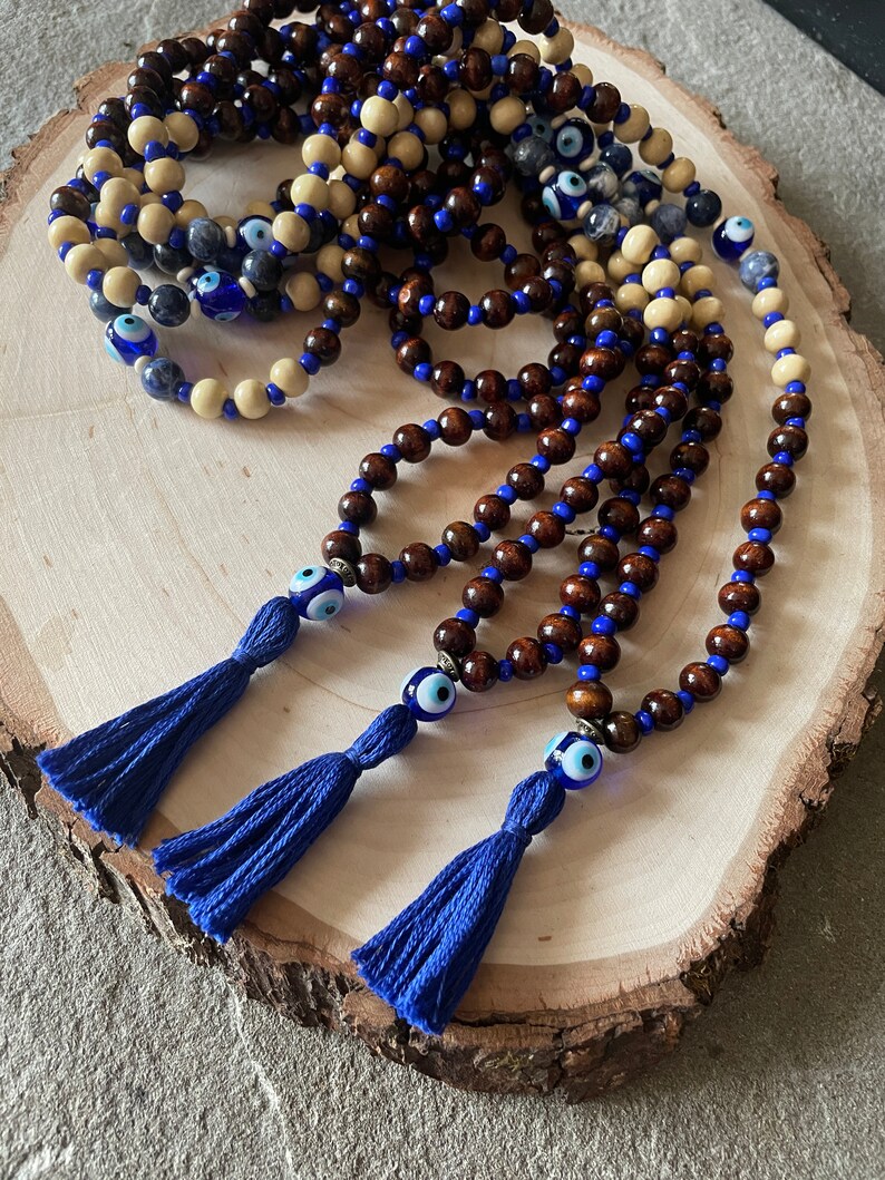 May include: A collection of beaded necklaces featuring wooden beads in brown and cream, with blue accents and tassels. Each necklace has an evil eye charm. The necklaces are presented on a wooden surface.