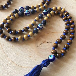 May include: A beaded necklace with wooden beads in brown and beige, blue beads, and eye-shaped glass beads. A blue tassel is attached. The necklace is displayed on a light wood surface.