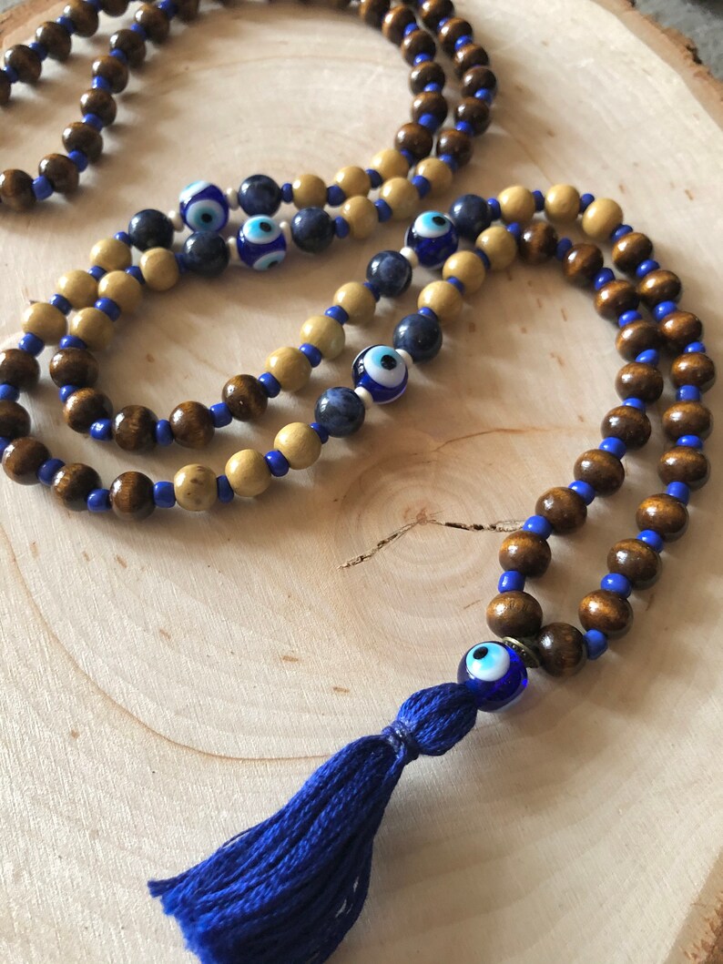 May include: A beaded necklace with wooden beads in brown and beige, blue beads, and eye-shaped glass beads. A blue tassel is attached. The necklace is displayed on a light wood surface.