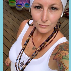 May include: A person wearing a white vest top and a white headscarf. They are wearing multiple beaded necklaces with charms and large hoop earrings. A colourful tattoo is visible on their arm. In the background are colourful singing bowls.
