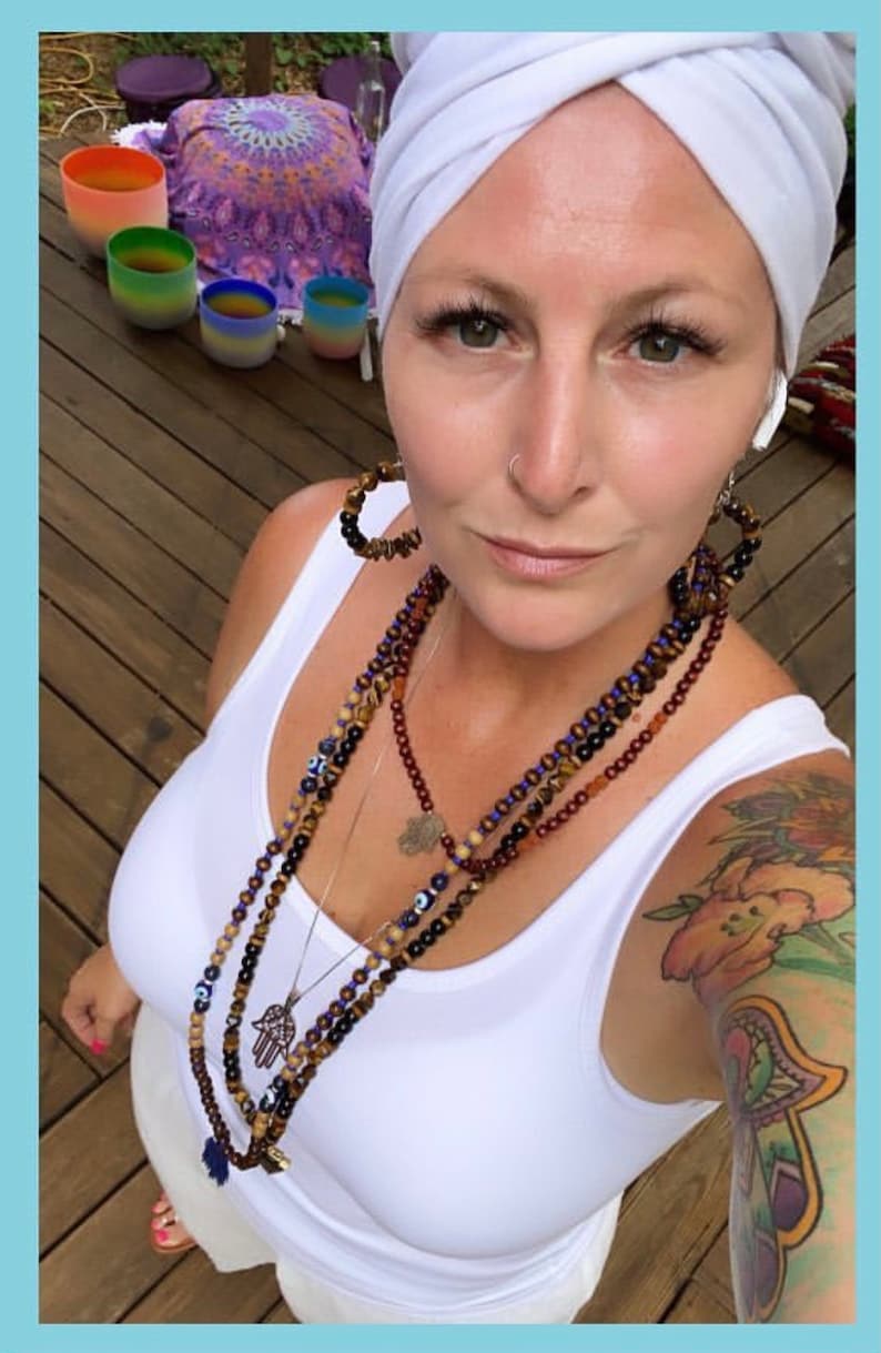 May include: A person wearing a white vest top and a white headscarf. They are wearing multiple beaded necklaces with charms and large hoop earrings. A colourful tattoo is visible on their arm. In the background are colourful singing bowls.