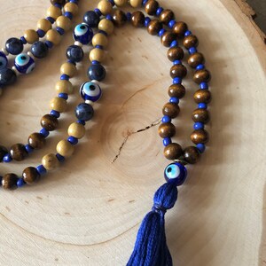 May include: Wooden beaded necklace featuring dark brown and light tan beads, with blue beads and blue evil eye beads. A blue tassel hangs from the bottom. The necklace is displayed on a wooden surface.