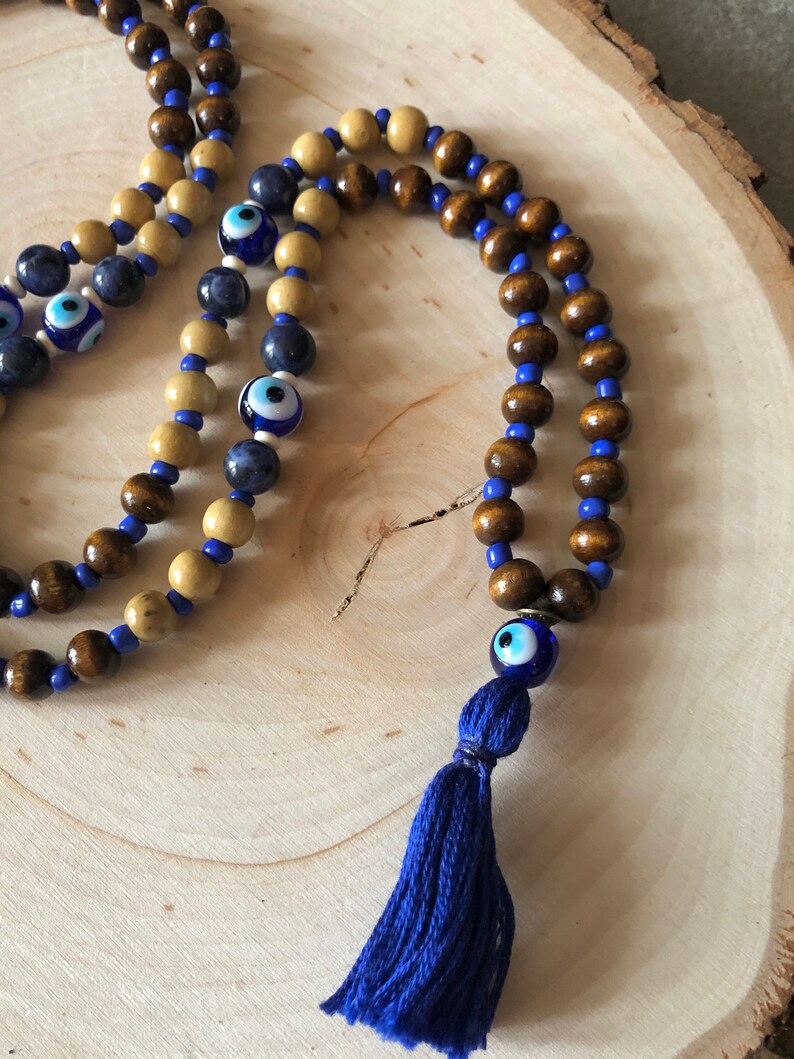 May include: Wooden beaded necklace featuring dark brown and light tan beads, with blue beads and blue evil eye beads. A blue tassel hangs from the bottom. The necklace is displayed on a wooden surface.