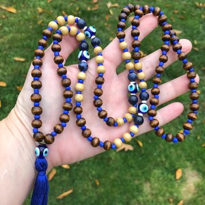May include: A beaded necklace with alternating dark brown and light wood beads, strung on blue cord. The necklace incorporates blue glass beads and eye-shaped beads. A blue tassel is attached at the end.