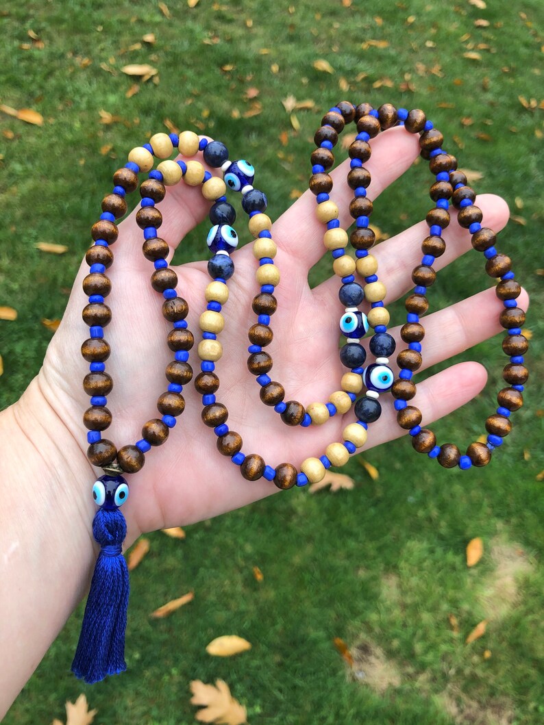 May include: A beaded necklace with alternating dark brown and light wood beads, strung on blue cord. The necklace incorporates blue glass beads and eye-shaped beads. A blue tassel is attached at the end.