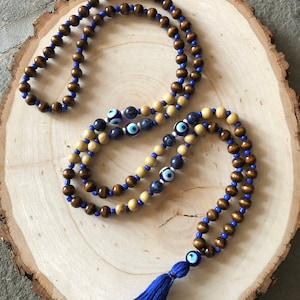 May include: Two beaded necklaces with brown and light tan wooden beads, strung on blue cord. One necklace features blue and white evil eye beads. A blue tassel with an evil eye bead hangs from one necklace. The necklaces rest on a wooden slice.