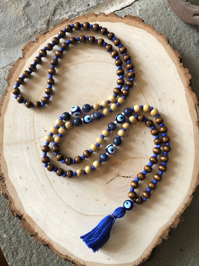 May include: Two beaded necklaces with brown and light tan wooden beads, strung on blue cord. One necklace features blue and white evil eye beads. A blue tassel with an evil eye bead hangs from one necklace. The necklaces rest on a wooden slice.