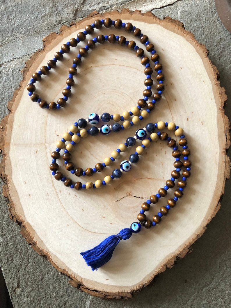 May include: A wooden bead necklace with a blue tassel. The necklace features alternating dark brown and light tan beads, connected by blue string. Some beads have an evil eye design.