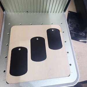 Op de afbeelding: Drie zwarte metalen hondenhangers op een licht houten oppervlak. De hondenhangers zijn rechthoekig met afgeronde hoeken en hebben een klein gaatje bovenaan.
