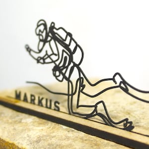 Könnte beinhalten: Schwarze Metallfigur eines Tauchers mit Taucherausrüstung, montiert auf einem Holzsockel. Der Name "MARKUS" ist in die Basis gelasert. Die Figur ist vor einem neutralen Hintergrund platziert.