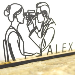 May include: A black laser-cut wooden decoration depicting a person looking through a camera at another person. The word "ALEX" is cut out below the figures. The figures are on a rectangular wooden base.