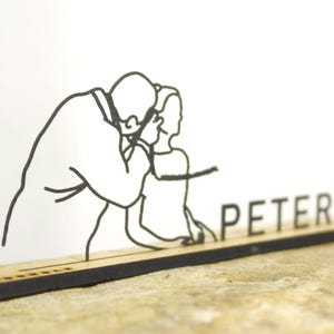 Op de afbeelding: Zwart-witte lijntekening van een persoon die het oor van een ander onderzoekt, met de naam "PETER" in zwarte letters op een houten voet. De voet is rechthoekig en rust op een stenen oppervlak.