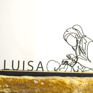 Può includere: Una scultura a filo nero di un professionista medico che esamina un bambino, con il nome "LUISA" davanti. La scultura è montata su una base di legno scuro, su sfondo bianco.