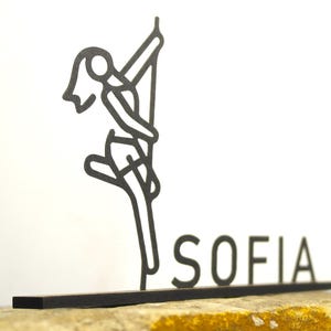 Op de afbeelding: Zwarte metalen sculptuur van een persoon die paaldanst, met de naam "SOFIA" in blokletters. De sculptuur is gemonteerd op een zwarte basis en tegen een witte achtergrond.